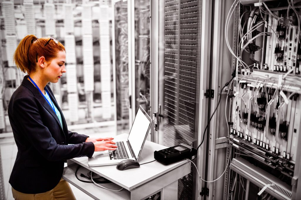 The image shows a female technician working in a data center. She is standing at a desk, using a laptop while analyzing or troubleshooting a nearby server rack. The server rack is filled with various equipment, cables, and components, indicating a complex setup. The technician is wearing a dark blazer and has a lanyard around her neck, suggesting a professional IT or engineering role. The background is mostly monochromatic, highlighting the technician and the laptop as the main focus of the image.