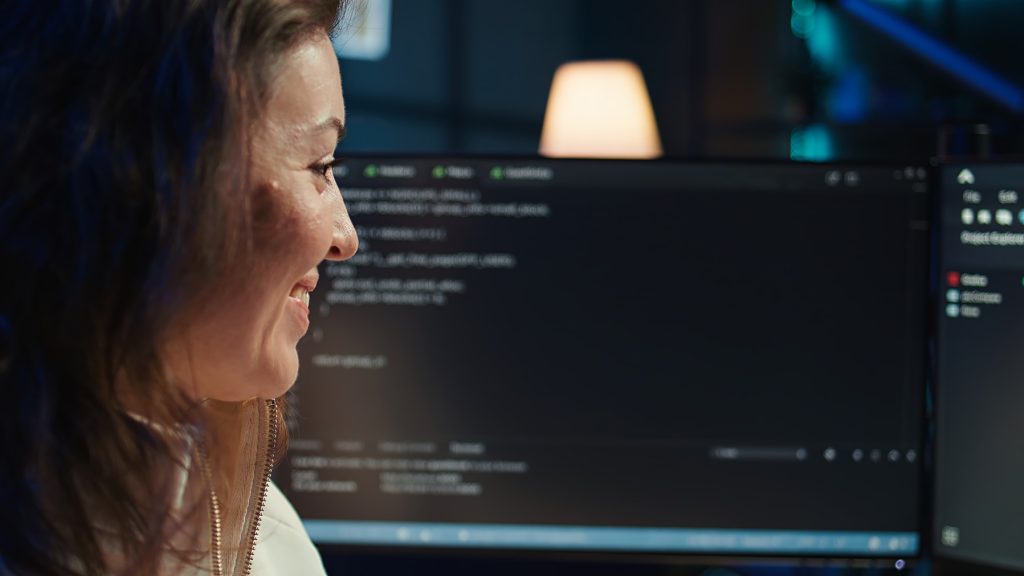 A woman smiling while looking at computer code on a screen in a dark workspace.