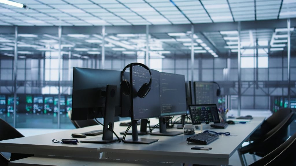 Modern computer workstation setup in a high-tech server room, featuring multiple monitors displaying code and system interfaces, with headsets hanging on the screens and various tech accessories like keyboards, a tablet, and a flash drive on the desk.