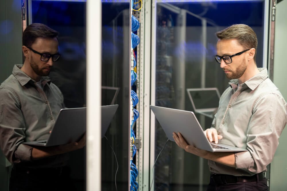 A man using a laptop in a server room, standing beside a glass cabinet reflecting his image. He is wearing glasses and a grey shirt, focused on working with the server equipment.