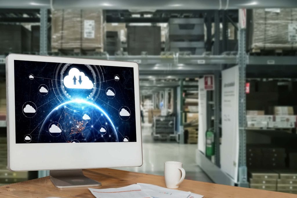 A computer monitor displaying a cloud computing network interface with interconnected cloud icons and a glowing Earth, placed on a desk in a warehouse filled with shelves and boxes, symbolizing cloud-based inventory or logistics management.
